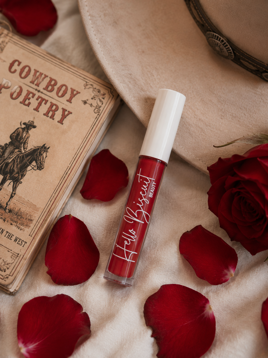 A clear tube of Hello Biscuit Beauty Two Dozen Roses red lip gloss on a textured surface surrounded by red rose petals, a cowboy hat, and a book titled Cowboy Poetry
