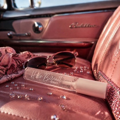 clear tube of Hello Biscuit Beauty Cowboy Casanova Clear Lip Gloss displayed on a plush pink leather vintage car seat next to sunglasses scattered pink crystals and a silk scarf