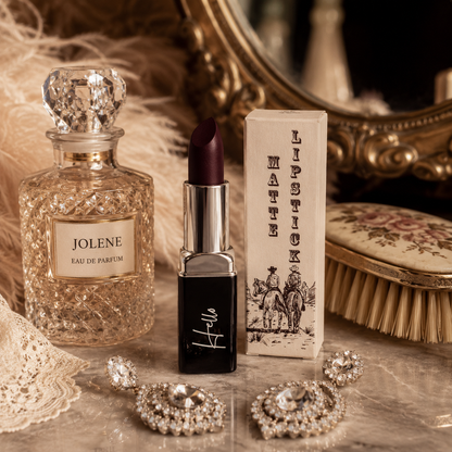 An open tube of Hello Biscuit Beauty Jolene matte lipstick standing next to its custom western box, a crystal Jolene perfume bottle, vintage lace, and sparkling jewelry on a rustic surface