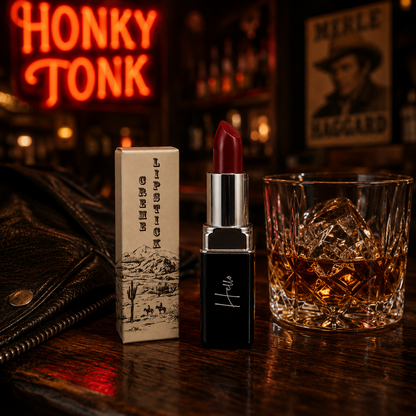 An open tube of Hello Biscuit Beauty Mamma Tried creme lipstick and its custom box on a wooden bar counter next to a glass of whiskey with a glowing red Honky Tonk neon sign in the background