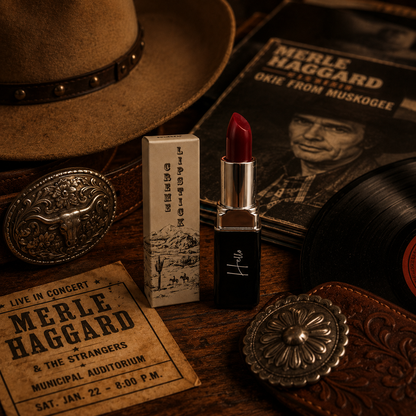 An open tube of Hello Biscuit Beauty Mamma Tried creme lipstick and its custom box on a rustic wooden table next to a Merle Haggard vinyl record album and a vintage concert ticket