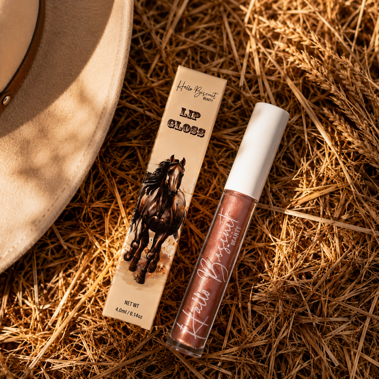A clear tube of Hello Biscuit Beauty Palomino Sparkly Mocha Lip Gloss and custom horse packaging resting on a rustic golden straw background next to a tan cowboy hat