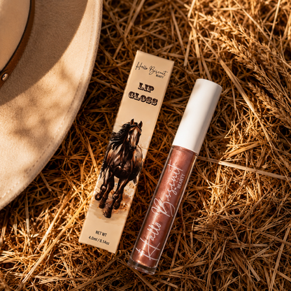 A clear tube of Hello Biscuit Beauty Palomino Sparkly Mocha Lip Gloss and custom horse packaging resting on a rustic golden straw background next to a tan cowboy hat