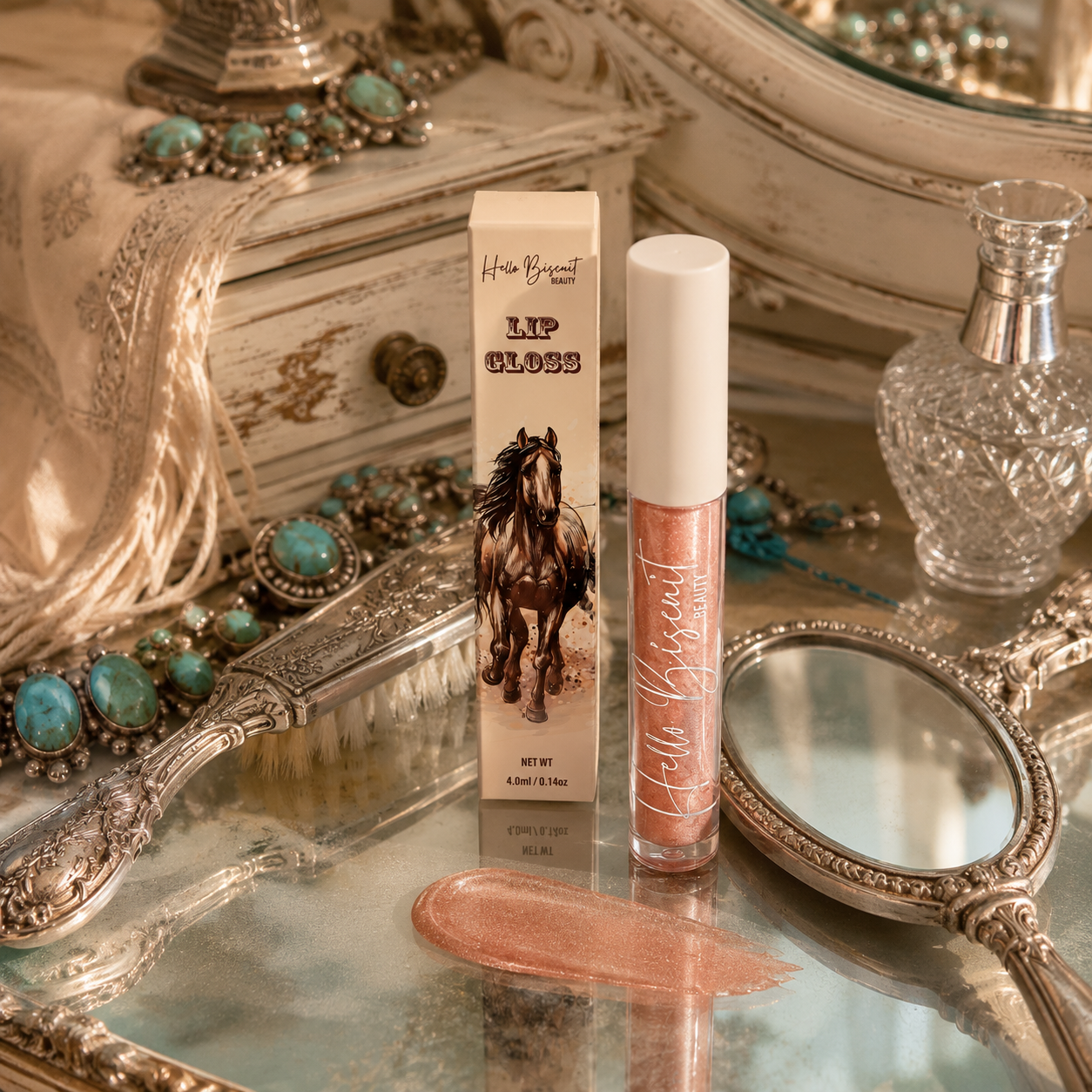 A clear tube of Hello Biscuit Beauty Palomino Sparkly Mocha Lip Gloss and horse design packaging box styled on a silver vintage vanity mirror next to a turquoise bracelet and a liquid makeup swatch