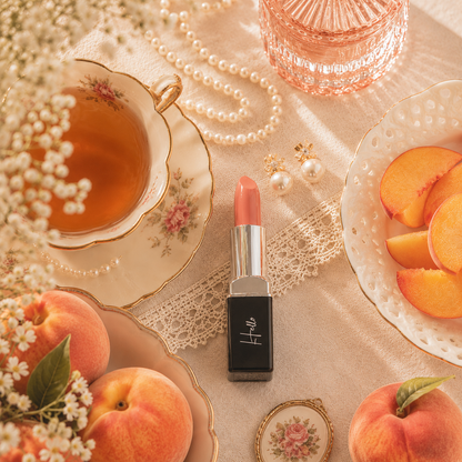An open tube of Hello Biscuit Beauty Peach Tea creme lipstick standing upright on a rustic surface surrounded by fresh peaches, a vintage teacup filled with tea, pearl jewelry, and an elegant crystal dish