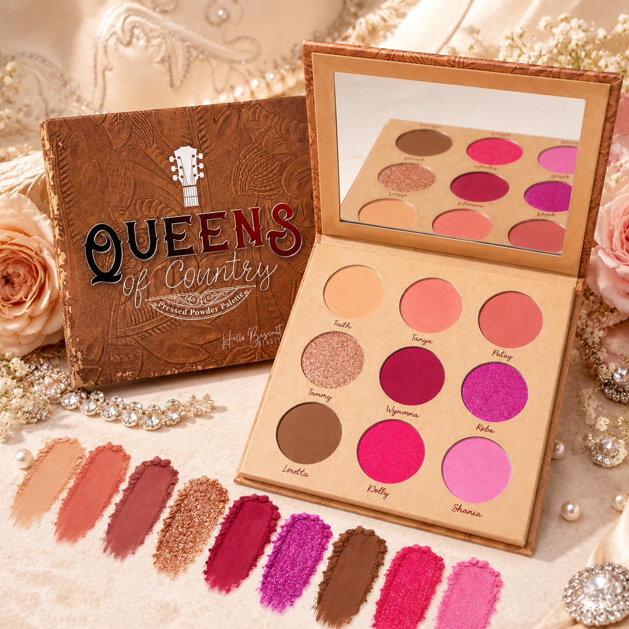 An open Hello Biscuit Beauty Queens of Country eyeshadow palette displaying nine matte and shimmer shades next to its closed brown embossed leather-style packaging on a rustic leather background with vibrant powder makeup swatches.